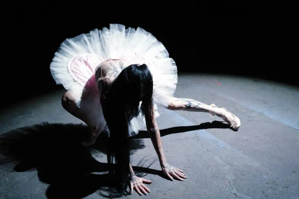 DeathbyRomy performing a dramatic, distorted ballerina pose in a pink tutu under a single spotlight, highlighting her signature "beauty in the macabre" style.