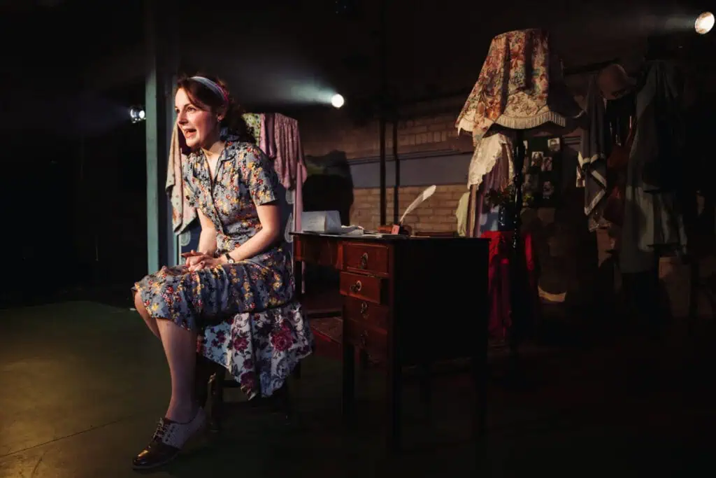 Actress Eva Feiler as Louise Rabiner sitting at a vintage wooden desk with a quill and letters in Ken Ludwig’s play Dear Jack, Dear Louise at Arcola Theatre.