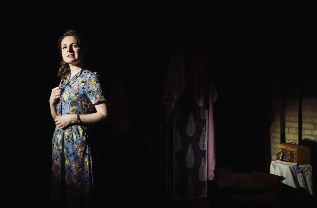 Eva Feiler performing as Louise in a 1940s floral dress, standing in a dramatic spotlight next to a vintage radio on stage.