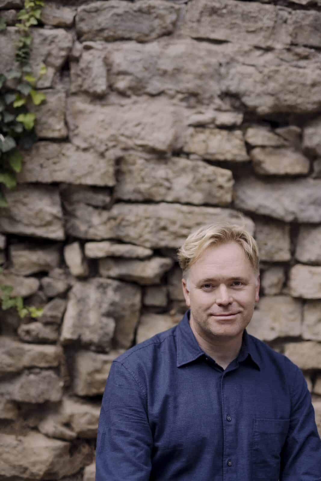 Graeme Richardson, author of the poetry collection Dirt Rich, posing in a blue button-down shirt in front of a rustic stone wall with green ivy.