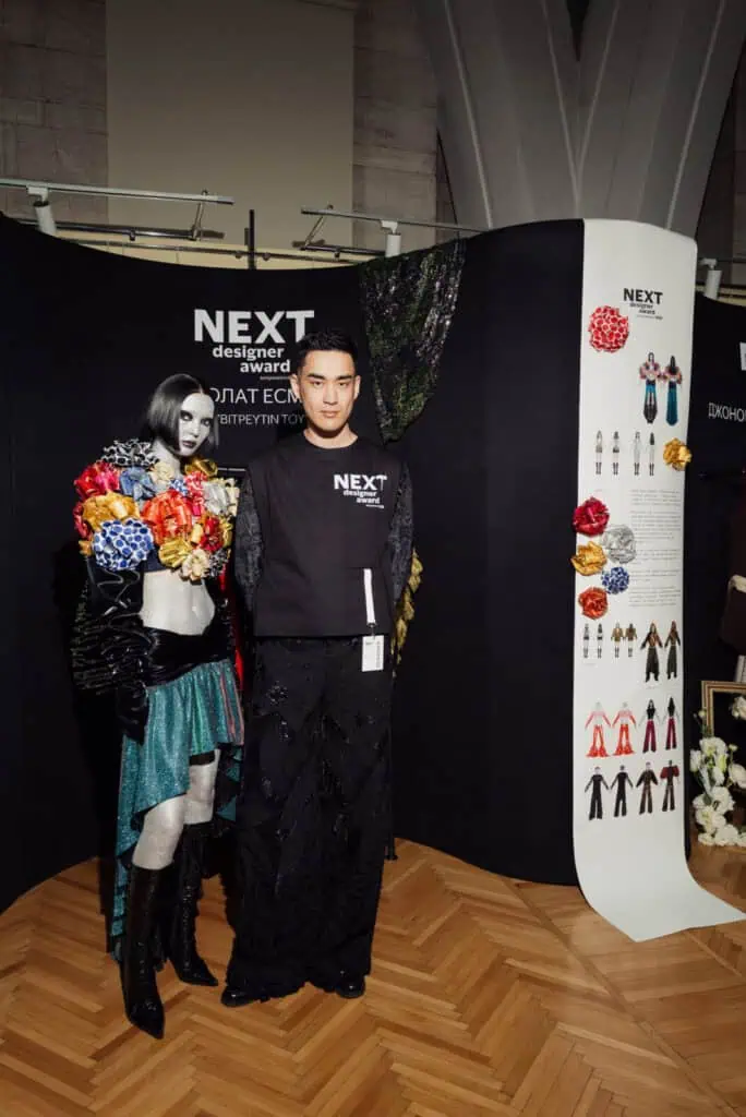 Winner Esbolat Esmukhanov standing next to his model in a booth at Visa Fashion Week Almaty, showcasing his award-winning designs that critique mass-market fashion culture.