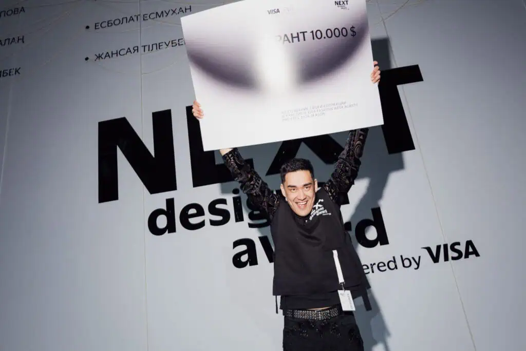 Designer Esbolat Esmukhanov holds up a $10,000 grant check at Visa Fashion Week Almaty after winning the NEXT Designer Award for his innovative "Fake Prada" collection.
