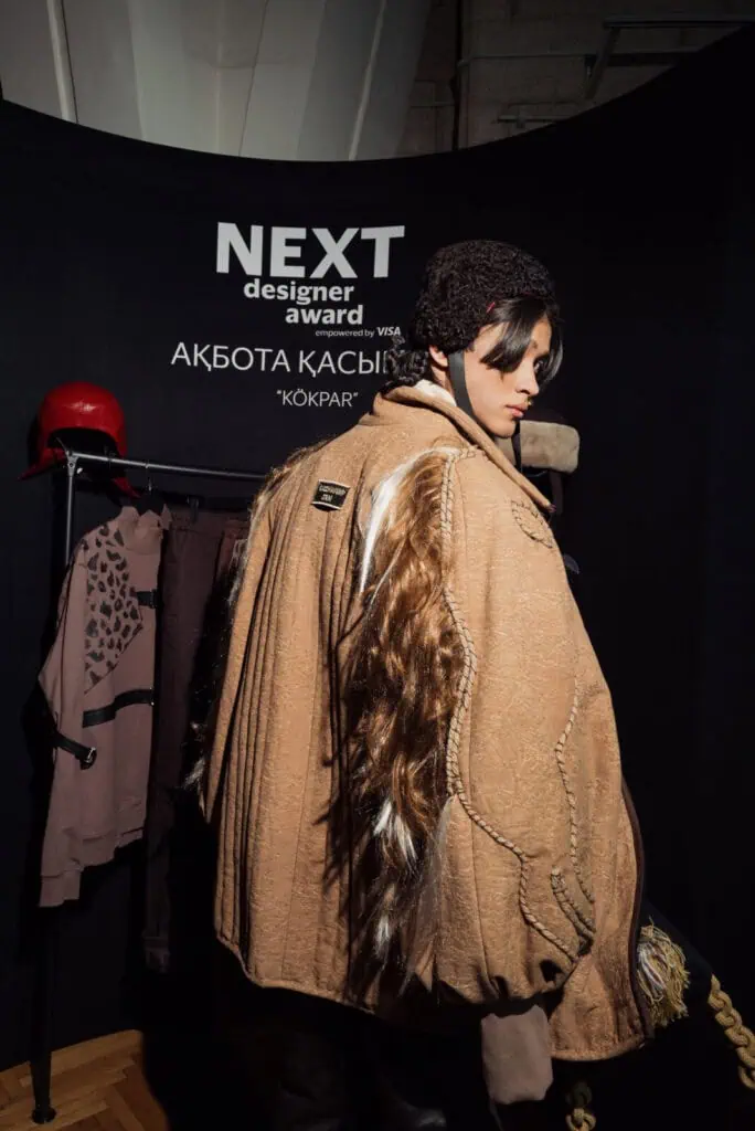 The back view of a tan quilted jacket by Akbota Kasymbek, featuring long faux fur trim and intricate braided rope details, symbolizing the strength of the Kazakh Steppe.