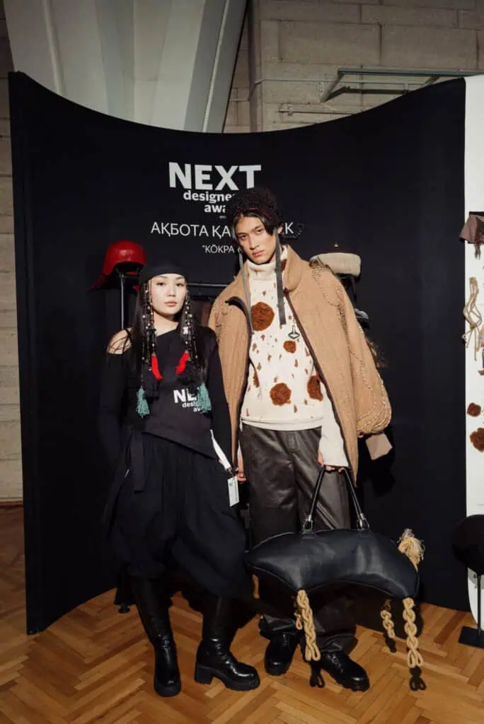 Designer Akbota Kasymbek posing with a model at Visa Fashion Week Almaty, featuring a look inspired by the traditional nomadic sport of Kökpar with an oversized textured coat and a horse-shaped leather bag.
