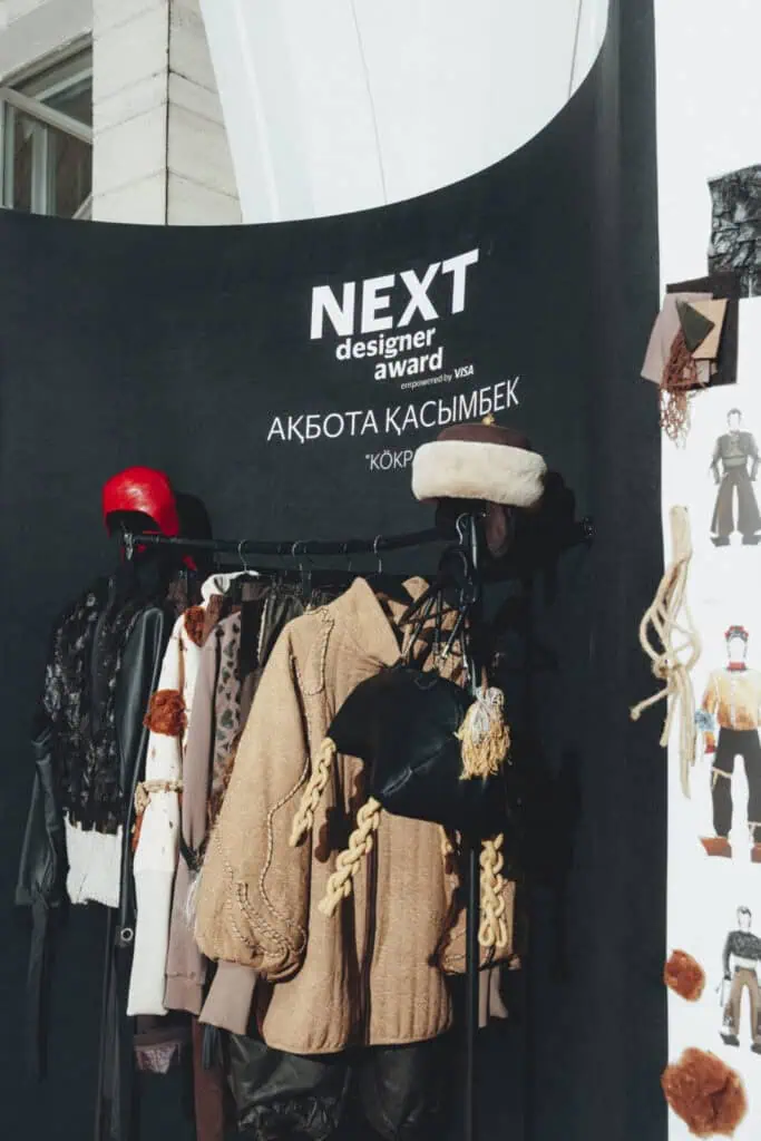The Akbota Kasymbek "Kökpar" exhibition booth at Almaty Fashion Week, displaying a rack of earth-toned garments, a red helmet, and a traditional fur hat.