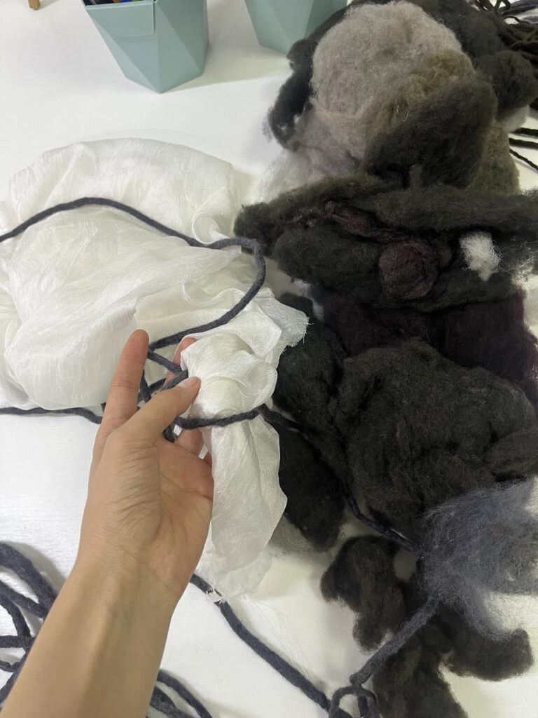 A close-up of a hand working with raw white silk and dark unspun wool fibers, demonstrating the artisanal felting process used for Meerim Naizabekova’s VFWA collection.