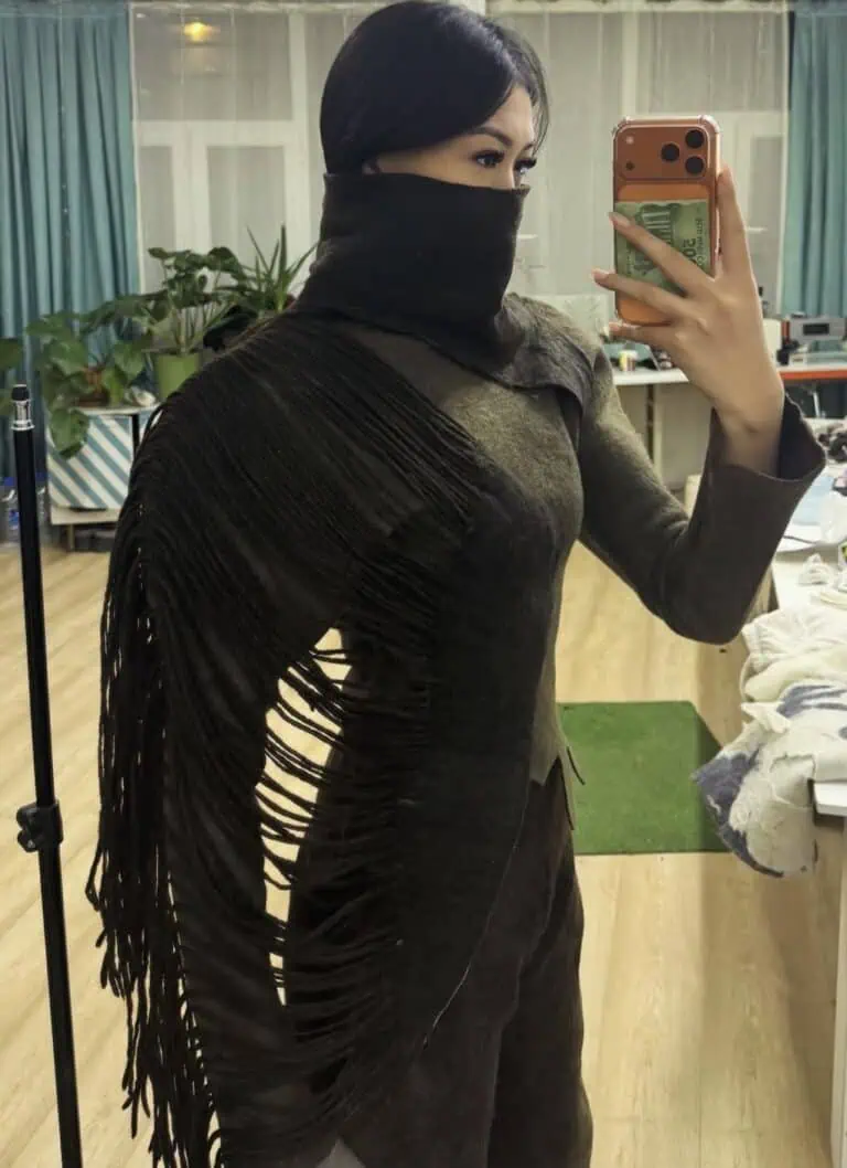 A close-up of a model wearing a dark, felted wool top by Meerim Naizabekova, featuring long, organic fringe and a built-in face covering as part of the "Balance" collection.