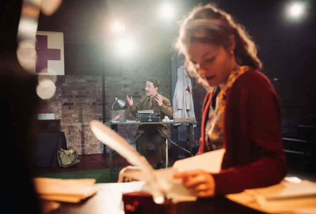 Preston Nyman using a vintage typewriter in a military office while Eva Feiler reads a letter in the foreground during a performance of Dear Jack, Dear Louise.