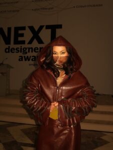 A close-up of a model wearing a maroon leather hooded jacket by Nuray Sakh, featuring rope-tie sleeve details and a face veil at Visa Fashion Week Almaty.