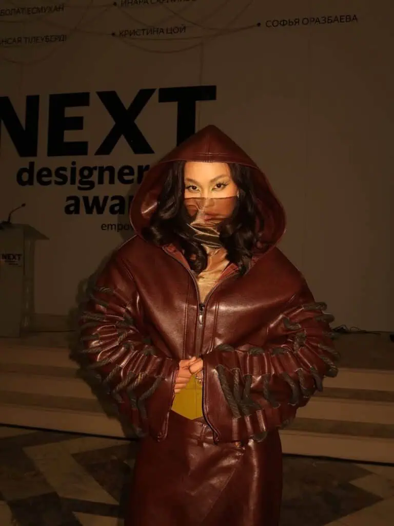 A close-up of a model wearing a maroon leather hooded jacket by Nuray Sakh, featuring rope-tie sleeve details and a face veil at Visa Fashion Week Almaty.