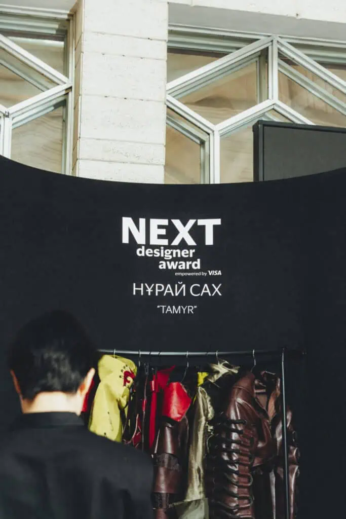 The NEXT Designer Award booth for Nuray Sakh's label SAKH, displaying the "Tamyr" collection against a black banner at Visa Fashion Week Almaty.