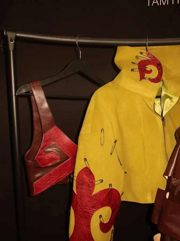 Close-up of a mustard yellow suede jacket by Nuray Sakh with red leather patches and safety pin details on a clothing rack during Almaty Fashion Week.