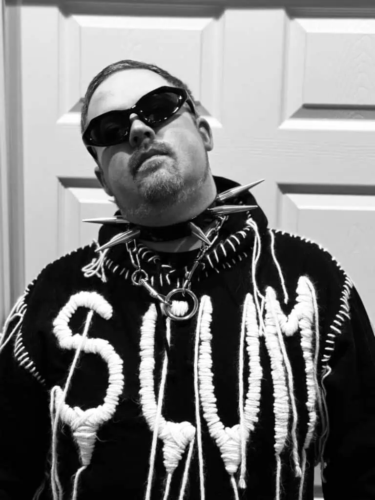 A black and white portrait of queer horror author Eric LaRocca wearing dark sunglasses and a spiked collar, embodying a bold and revolutionary aesthetic.