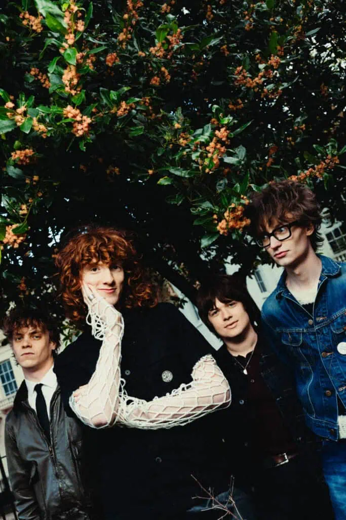 The four members of The Sukis standing beneath a flowering tree in bloom, one with red curly hair resting a hand on their cheek, another in a denim jacket with glasses — photograph by Nathan Cole