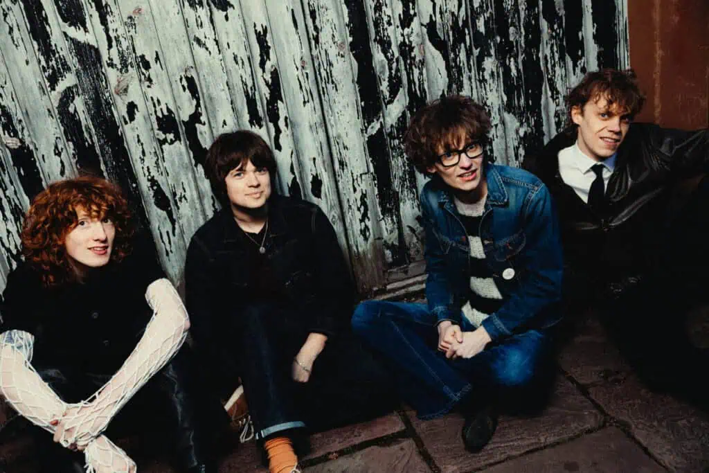 The four members of The Sukis sitting against a peeling painted wooden door, smiling candidly, dressed in denim, mesh sleeves, and a striped knit — photograph by Nathan Cole