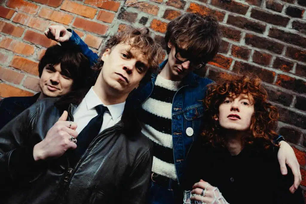 Close-up of The Sukis against a red brick wall, one member leaning forward with a raised fist, another in a white shirt and tie, a third in a striped jumper, and the fourth with red curly hair — photograph by Nathan Cole
