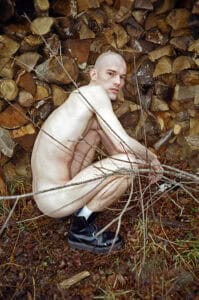 A shaved-head man crouching nude among bare branches in front of a stacked wood pile, wearing only white socks and black boots, looking directly into the camera — photograph by Slava Mogutin