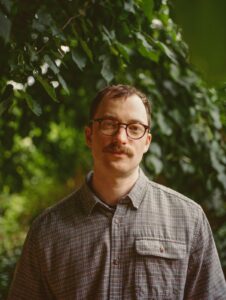 Author Jem Calder posing for a portrait in a gray plaid shirt and glasses against a lush green foliage background.