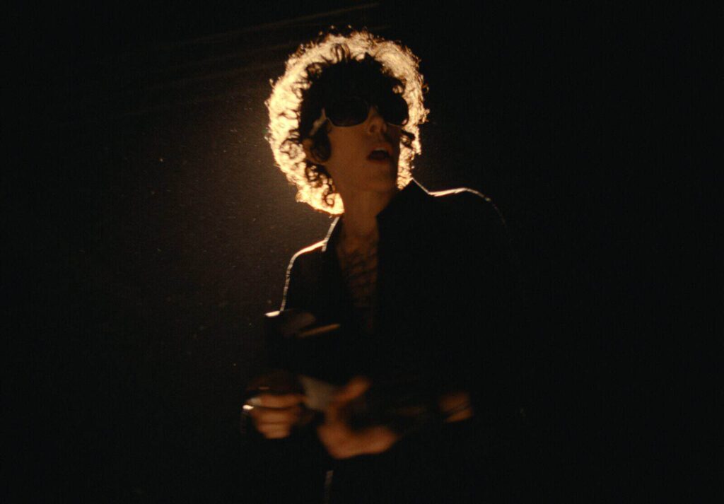 A dramatic low-light silhouette of artist LP, featuring their iconic curly hair and sunglasses highlighted by a strong backlight against a dark, hazy background.