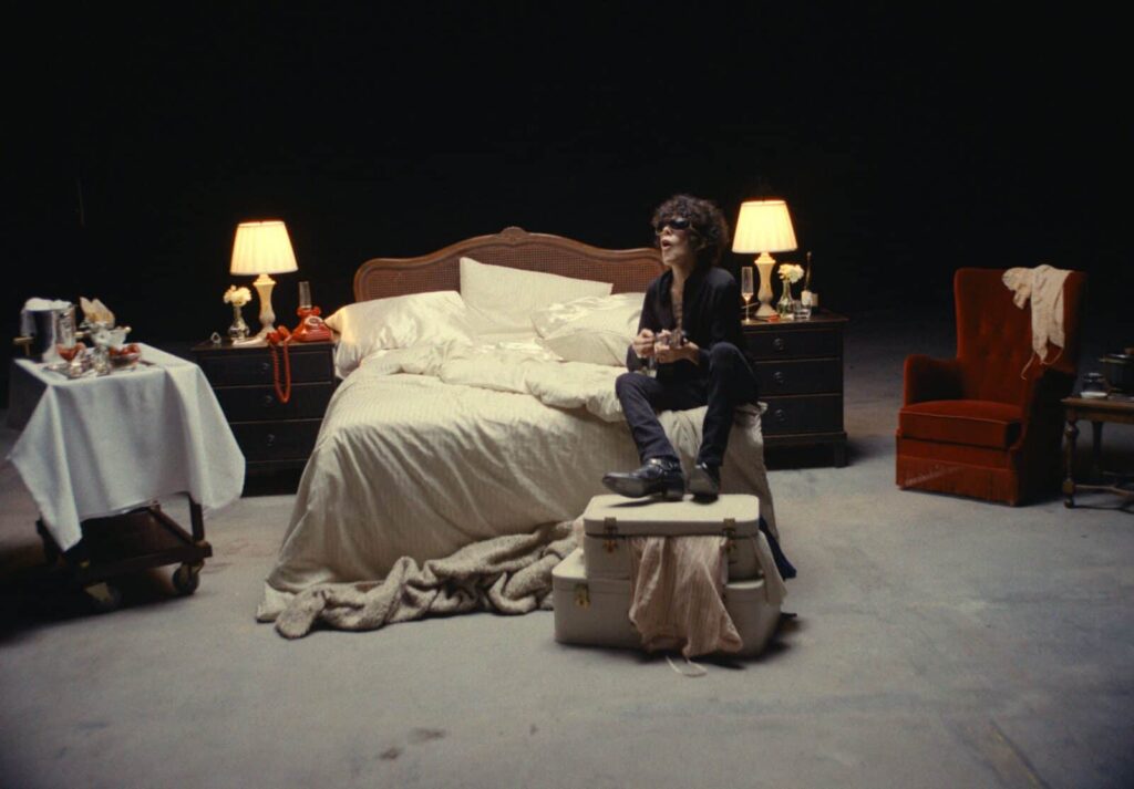 LP sitting on a vintage suitcase at the foot of an unmade bed on a dark music video set, surrounded by warm bedside lamps, a red velvet armchair, and a tray of champagne and strawberries.