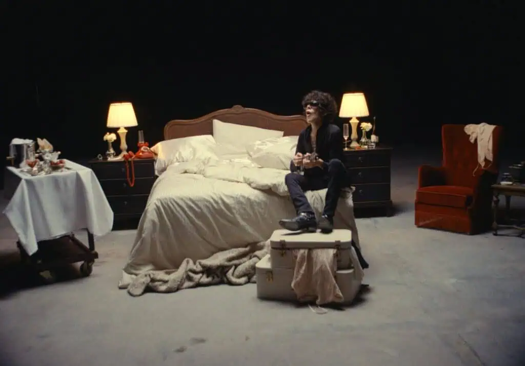 LP sitting on a vintage suitcase at the foot of an unmade bed on a dark music video set, surrounded by warm bedside lamps, a red velvet armchair, and a tray of champagne and strawberries.