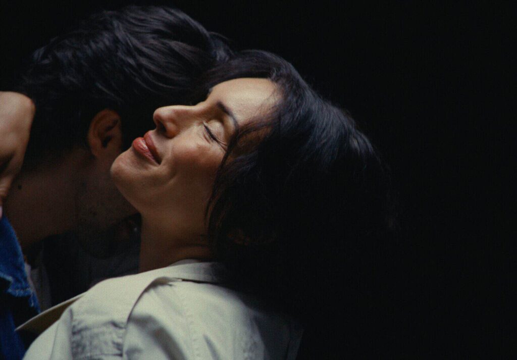 A warm, intimate close-up of a woman with her eyes closed and a peaceful smile, leaning into a man's embrace against a soft, dark backdrop in the new LP Shelly music video.