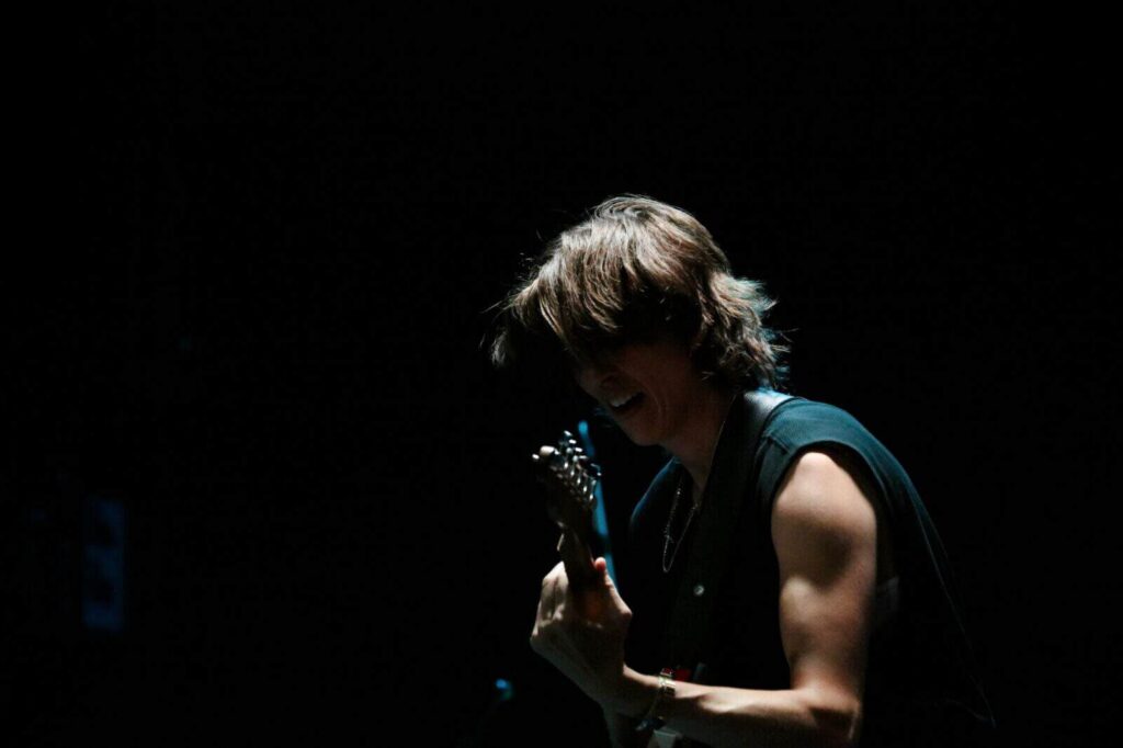 Side profile of E.R. Fightmaster playing electric guitar on a dark stage under a spotlight