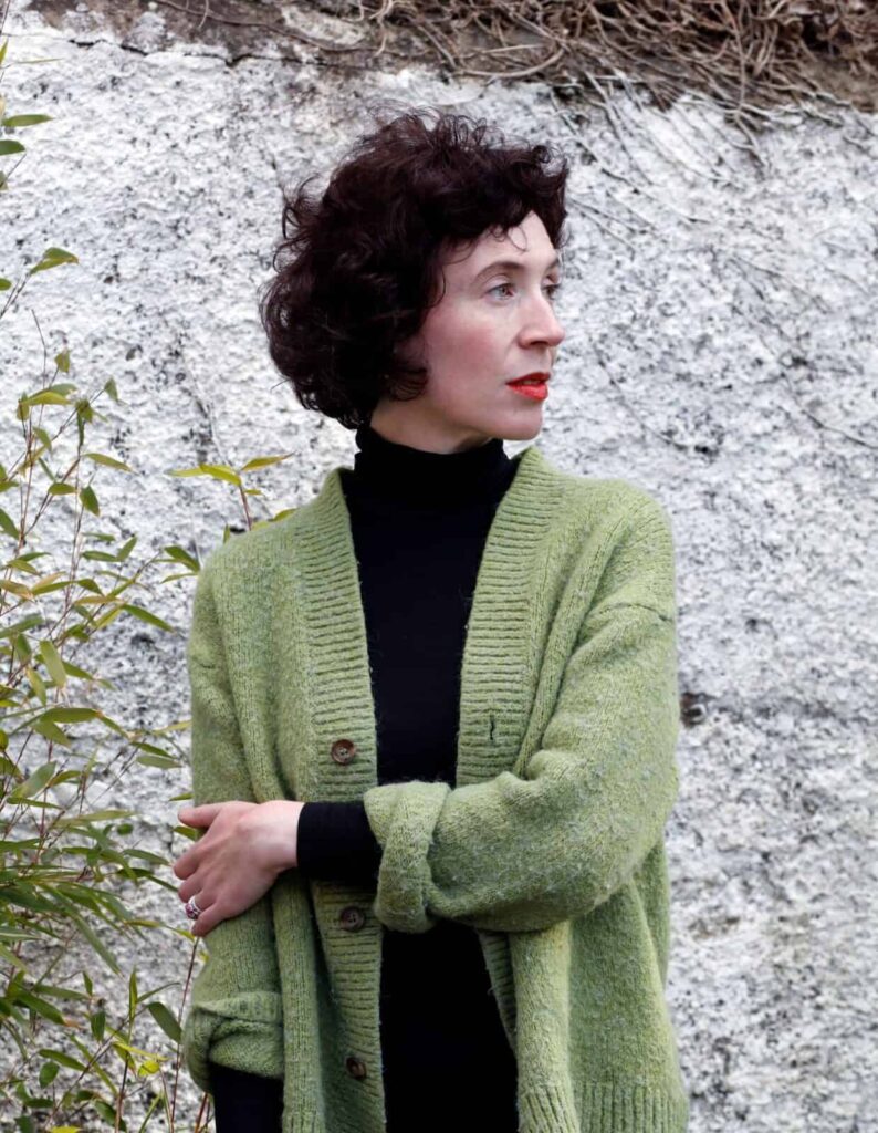 Niamh Campbell wearing a green cardigan and black turtleneck standing against a stone wall