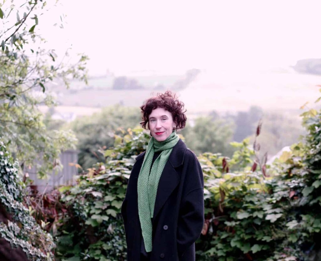 Niamh Campbell smiling in a black coat and green scarf with a countryside background