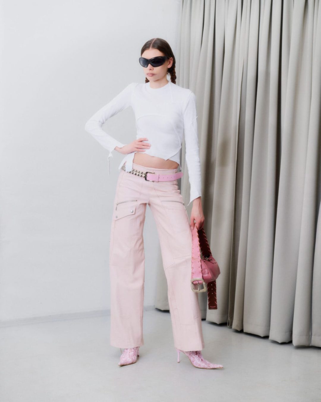 A person poses indoors wearing a fitted white long-sleeve top, light pink wide-leg pants with a pink belt, pink patterned boots, large black sunglasses, and carries a pink studded handbag. Beige curtains hang in the background.