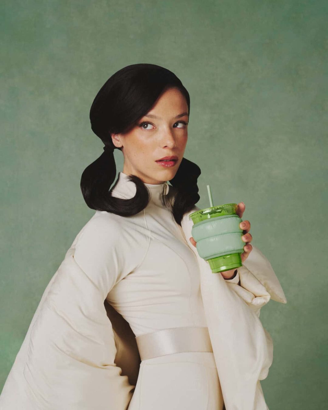 A woman with dark, styled hair and a serious expression wears a light, futuristic outfit and holds a green collapsible cup with a straw against a plain green background.