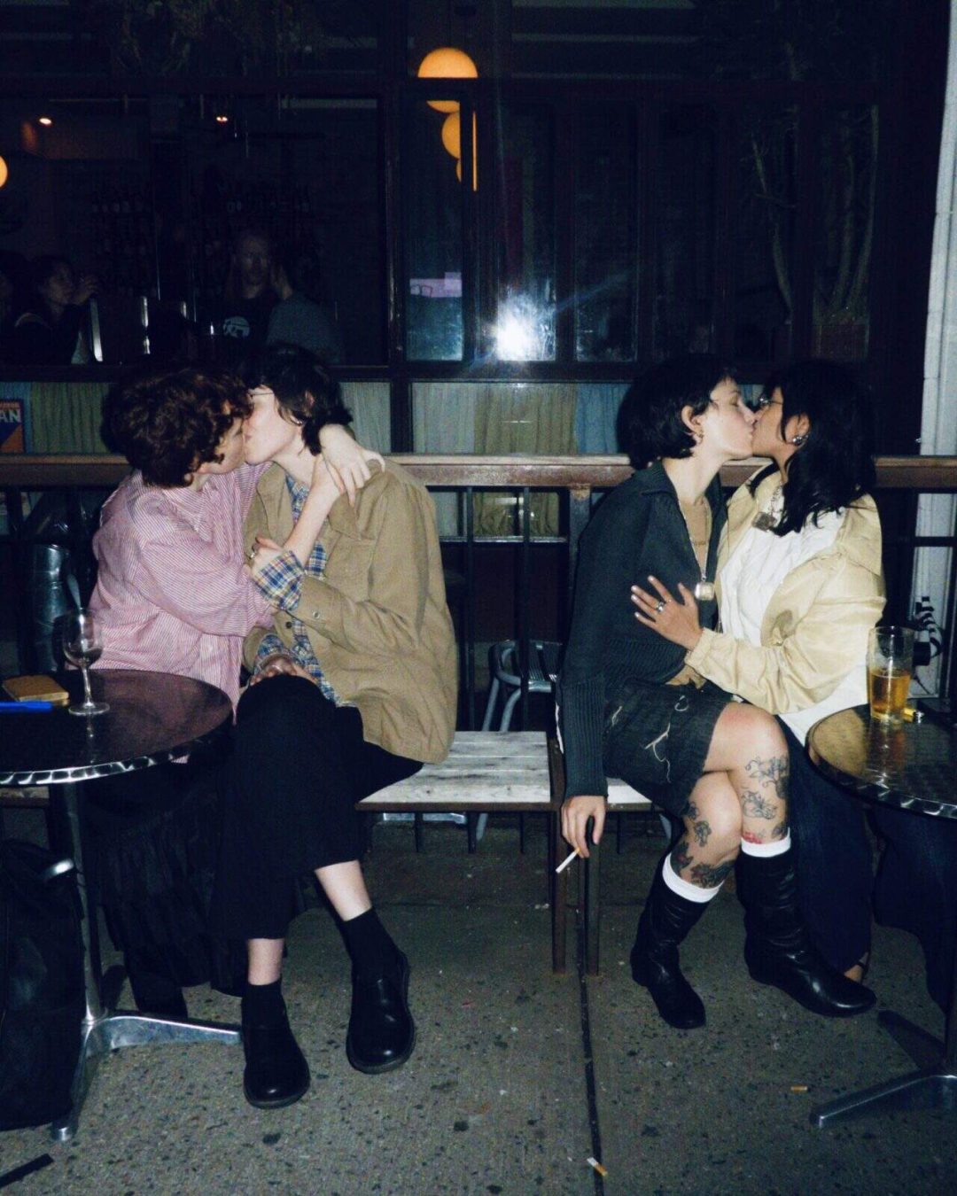 Two couples sit closely together on a bench outside a bar at night, each pair kissing. There are drinks on the tables beside them, and the scene has a warm, intimate, and candid atmosphere.