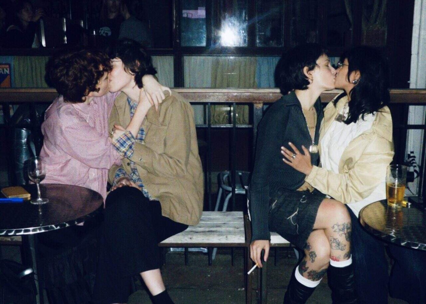 Two couples sit closely together on a bench outside a bar at night, each pair kissing. There are drinks on the tables beside them, and the scene has a warm, intimate, and candid atmosphere.