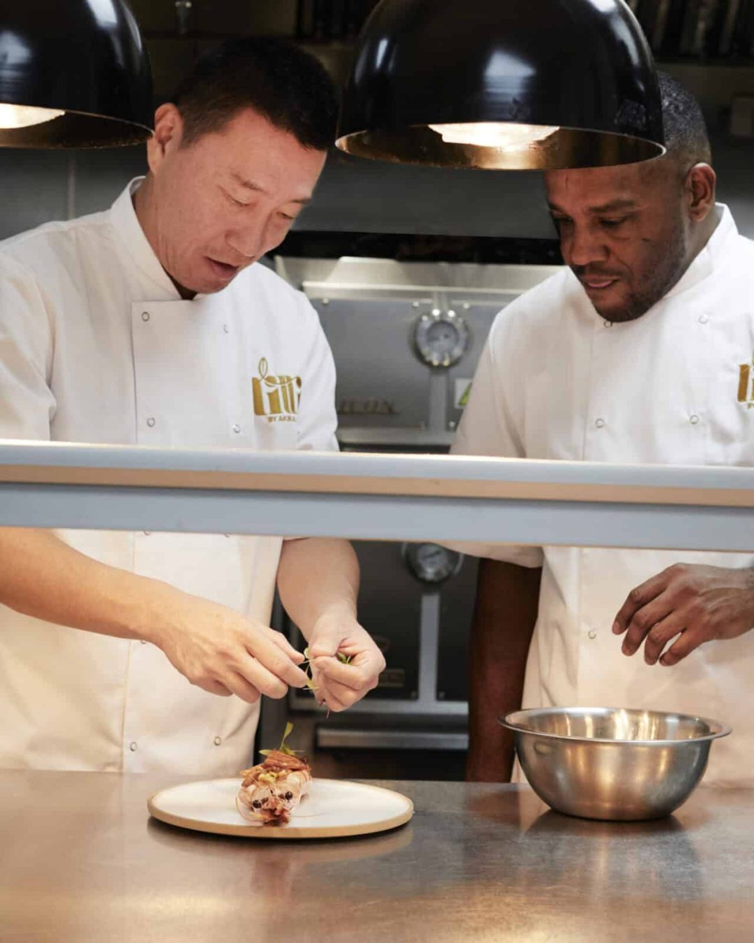 Two chefs in white uniforms prepare a dish together in a professional kitchen. One chef adds garnish to food on a plate, while the other watches, standing beside a metal mixing bowl.