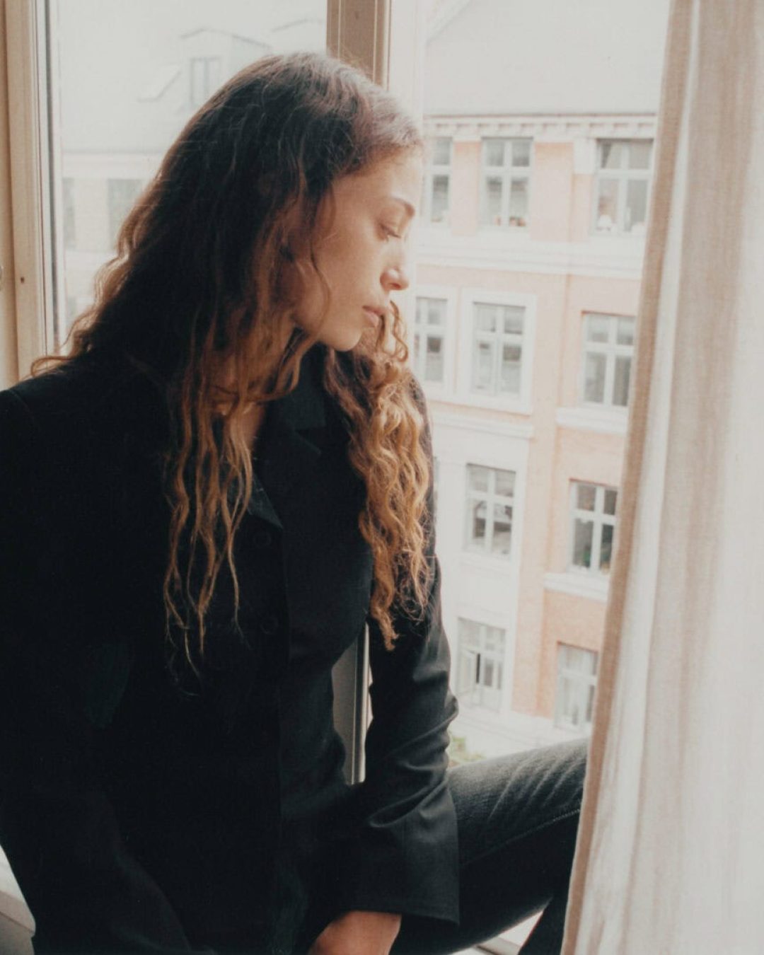 A medium shot of Alba Akvama sitting on a windowsill between soft, neutral curtains. She is wearing a black blazer and looking down pensively, with European-style brick architecture visible through the glass behind her.
