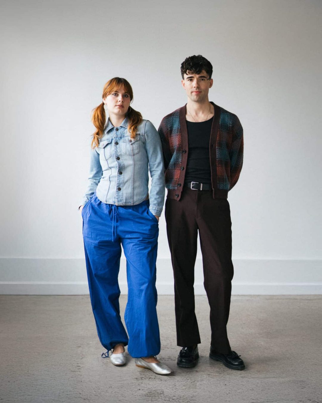 A woman in a denim jacket and loose blue pants stands next to a man wearing a patterned jacket over a black shirt and brown pants. Both face forward in a simple, light-filled room with plain walls.