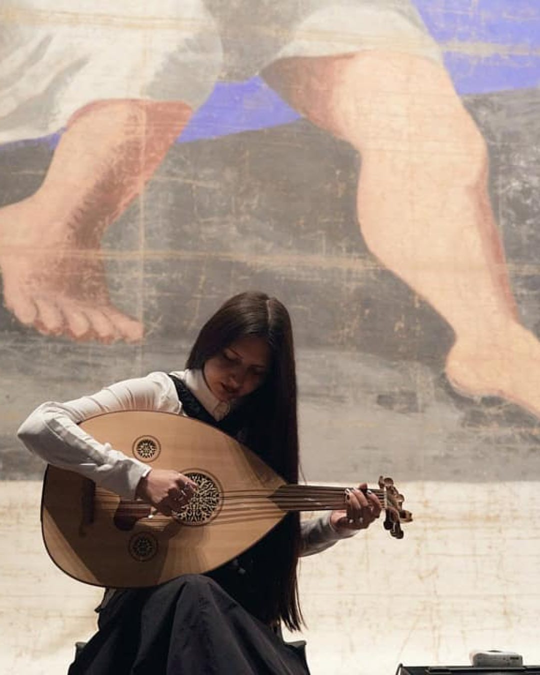 A wide shot of musician BODUR seated and playing a traditional oud. She is positioned in front of a large-scale mural, with a double bass visible to the right, highlighting her interdisciplinary musical style.