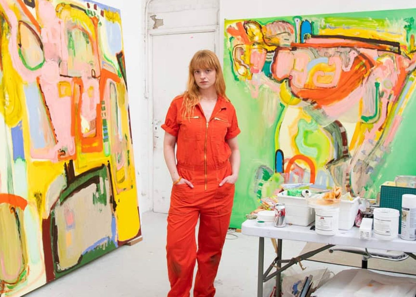 Portrait of artist Bunny Hennessey in a bright studio, standing between two large abstract canvases in vibrant colours, surrounded by paints and tools.