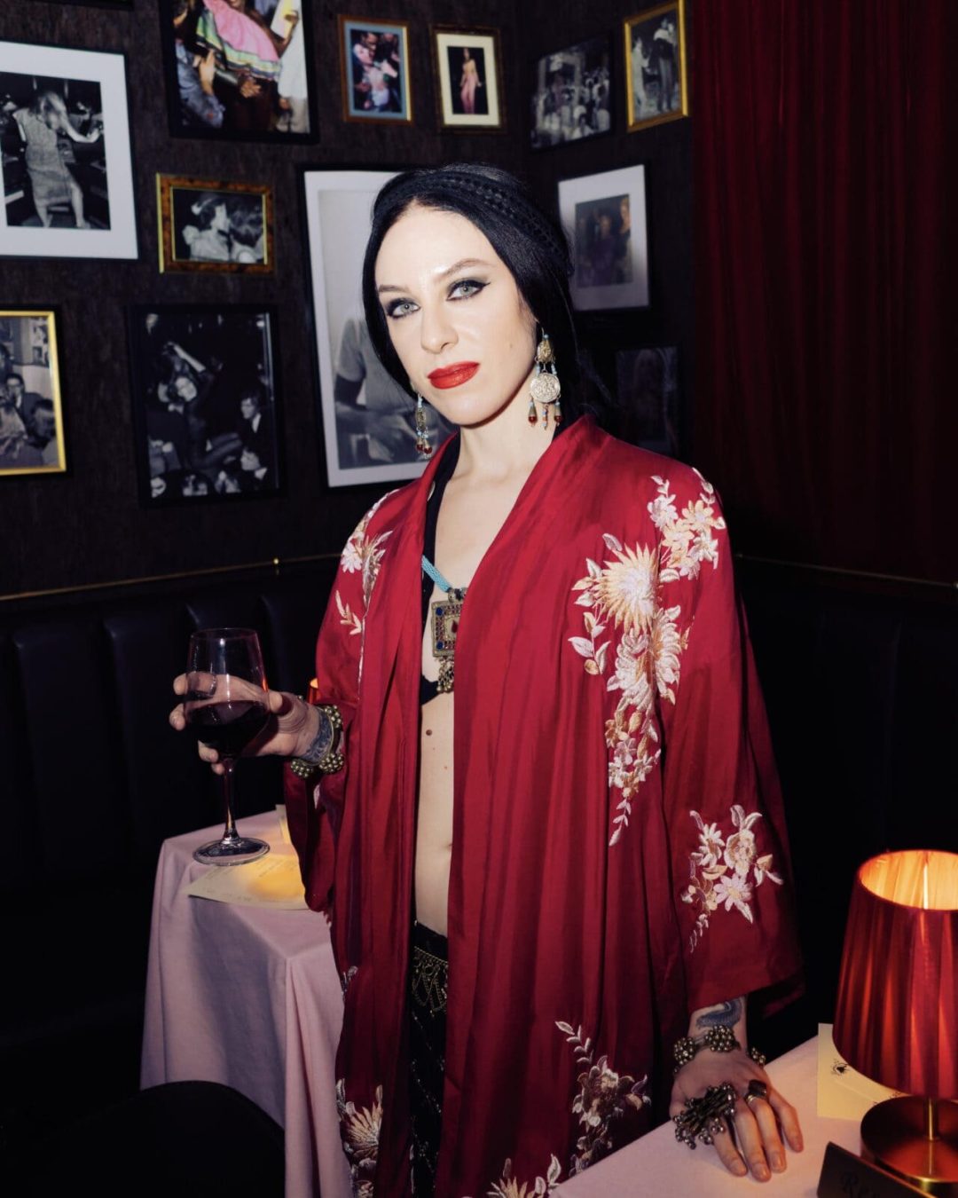 A person wearing a red floral robe and bold makeup stands indoors, holding a glass of wine. The setting is dimly lit with framed photos on the wall, red curtains, and a small table with a lamp in the background.