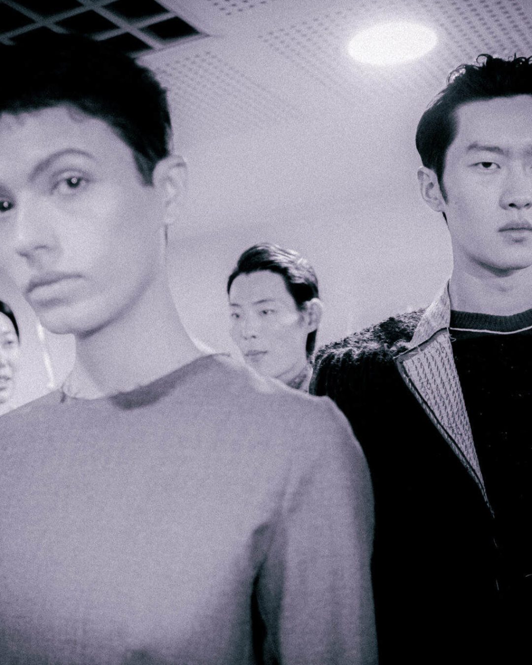 A group of four people stand indoors under bright ceiling lights, with two individuals in the foreground looking serious and two others behind them, slightly out of focus. The image has a muted, monochrome tone.