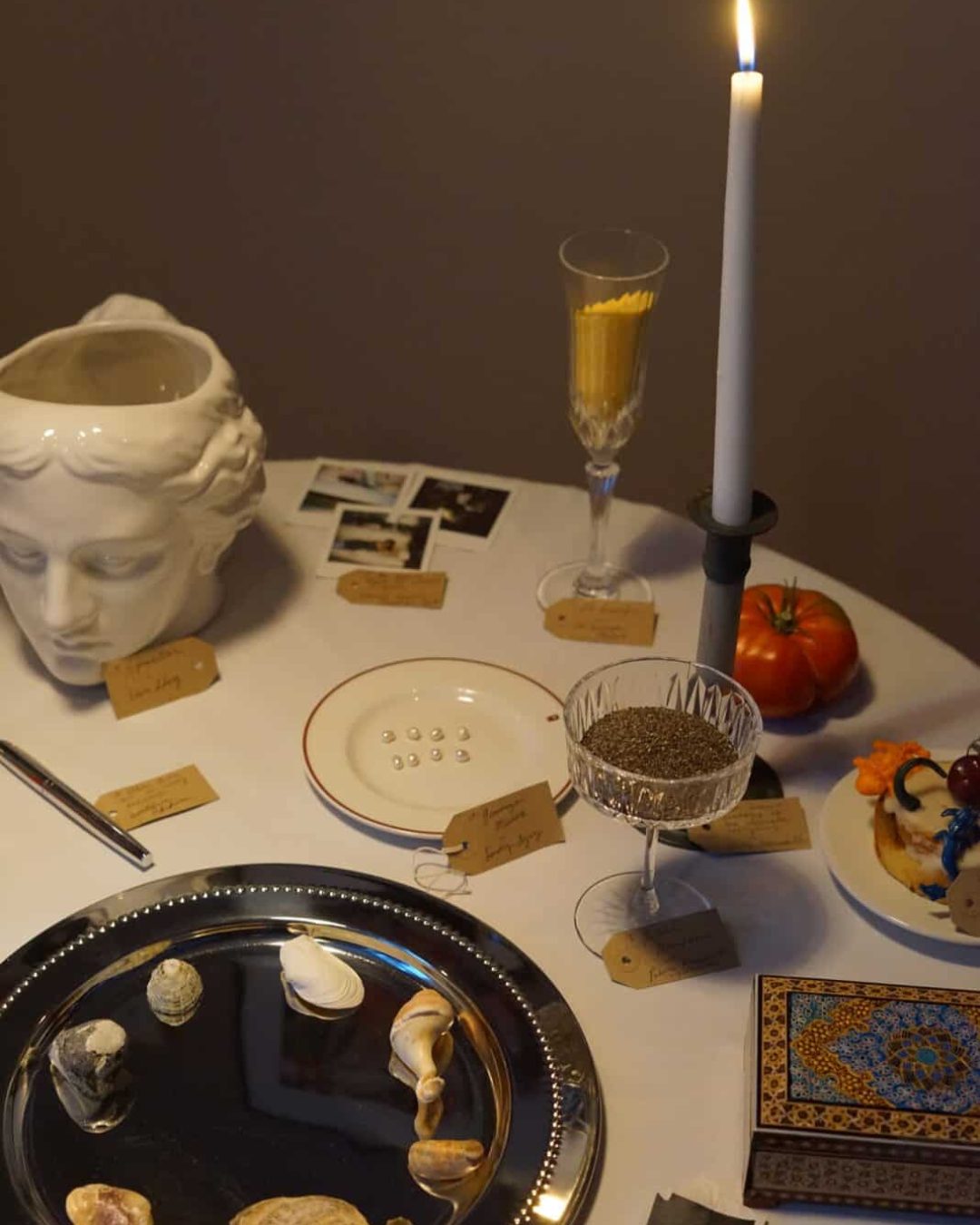 A round table at Perediza IV presentation displays seashells, a silver tray, a candle, a statue head, vintage photos, a plate with pills, labeled tags, a crystal glass with seeds, a tomato, and a slice of cake with flower decorations.