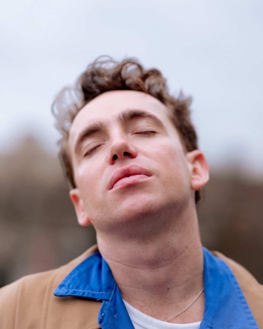 A person with short, curly brown hair and closed eyes tilts their head back slightly, appearing relaxed or contemplative. They wear a blue collared shirt under a tan jacket, with a blurred outdoor background.
