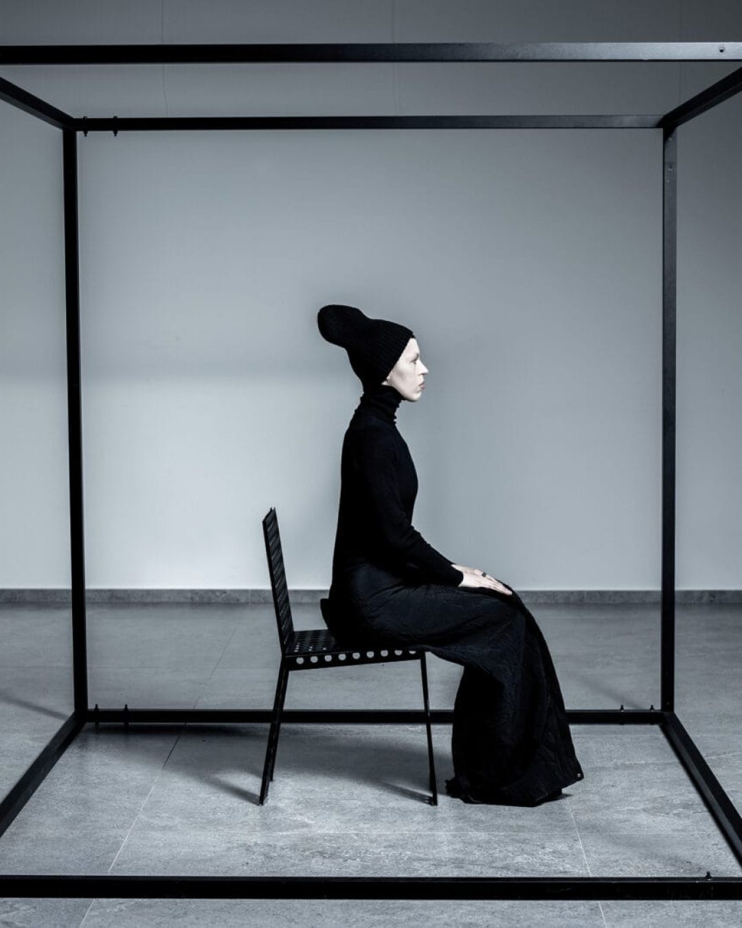 A person dressed in all black sits on a chair inside a large, open black metal cube frame in a minimalist, white room. The person is in profile, wearing a tall hat, and appears contemplative.