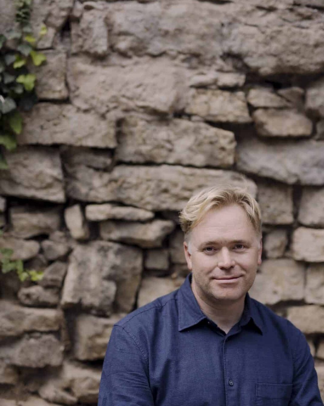 Graeme Richardson, author of the poetry collection Dirt Rich, posing in a blue button-down shirt in front of a rustic stone wall with green ivy.
