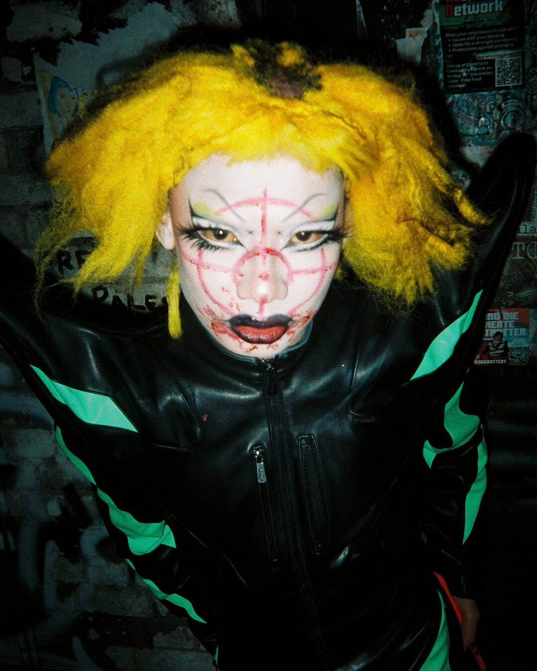 A person with messy bright yellow hair and dramatic clown-like makeup, including a red target pattern on their face, poses in a black and neon green outfit against a graffiti-covered wall at the Harringay Warehouses.