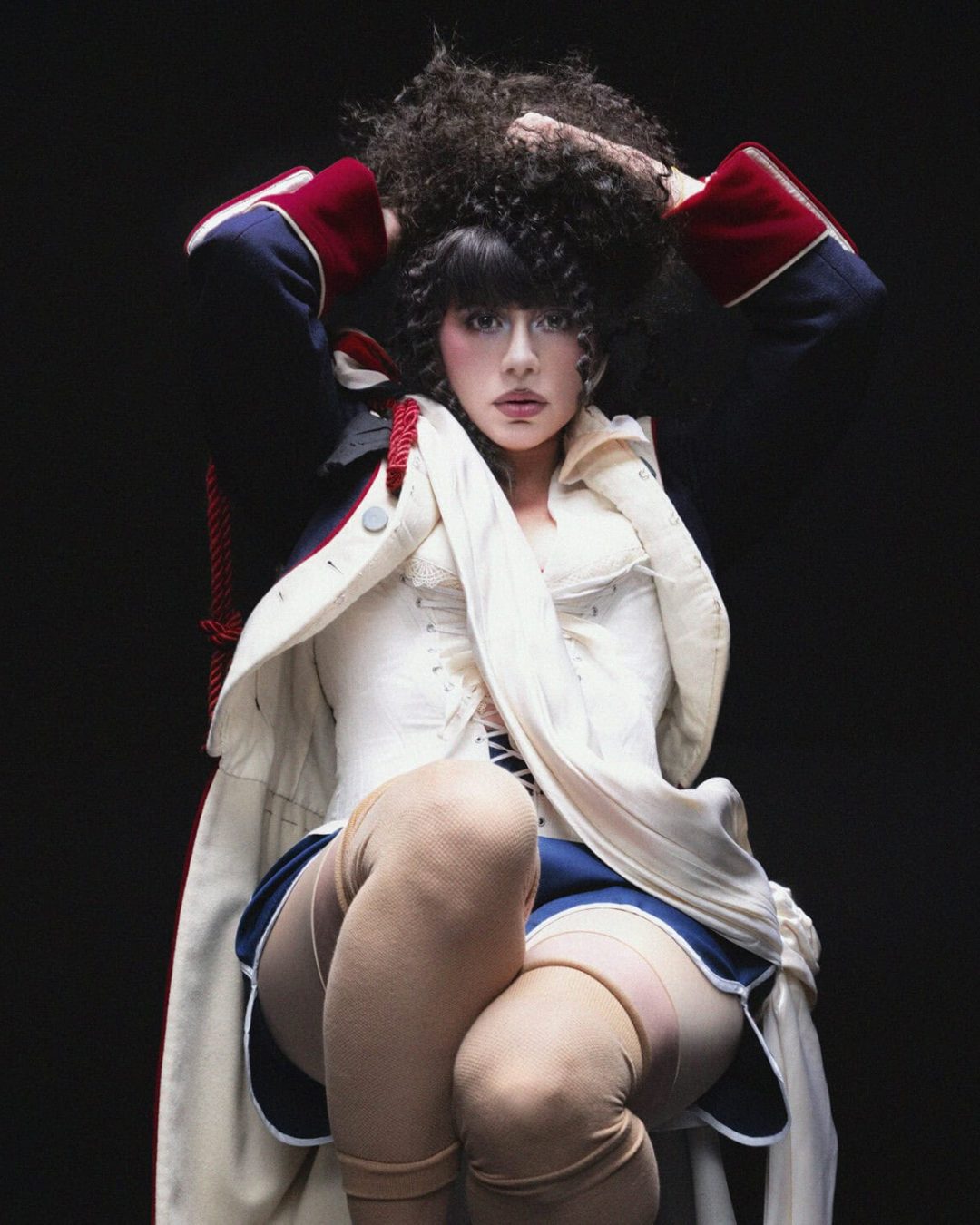 KÁRYYN in theatrical makeup and costume, wearing a dark jacket with red cuffs, a white corset, blue skirt, and beige stockings, sits with hands in curly hair against a dark background.