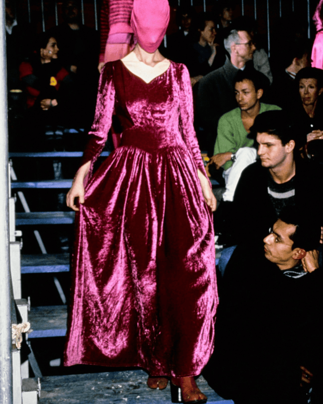 A model wearing a vibrant pink velvet gown reminiscent of Margiela AW95 and a matching pink face-covering walks down steps at a fashion show, with audience members seated closely on either side.