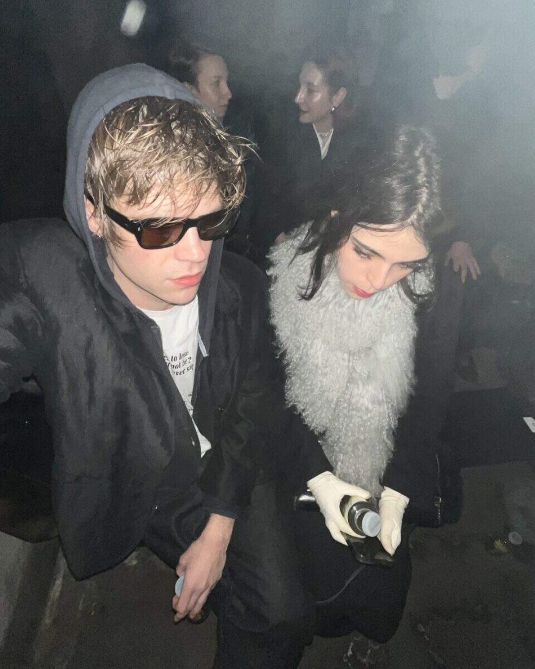 Two people sit close together in a dim, smoky setting. The person on the left wears sunglasses and a black hoodie, while the person on the right wears a fluffy scarf and white gloves, looking down at something in their hands. Other people are in the background.