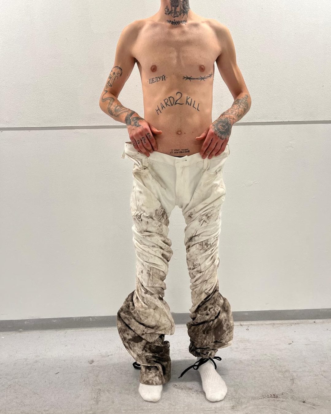 A shirtless person with tattoos on their chest, arms, and neck stands against a white wall —wearing loose, dirty white pants bunched by Kalina Dorożyńska Designs at the ankles and white socks; their hands rest on their hips.