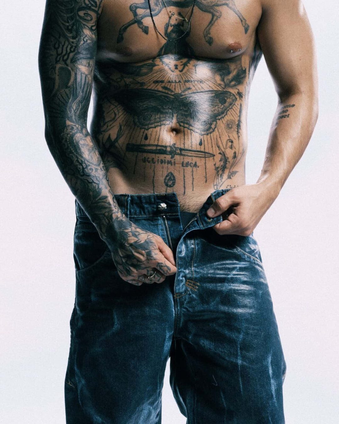 A shirtless person with extensive tattoos on their chest, arms, and torso is standing and pulling up blue jeans, partially unzipped, against a plain light background.